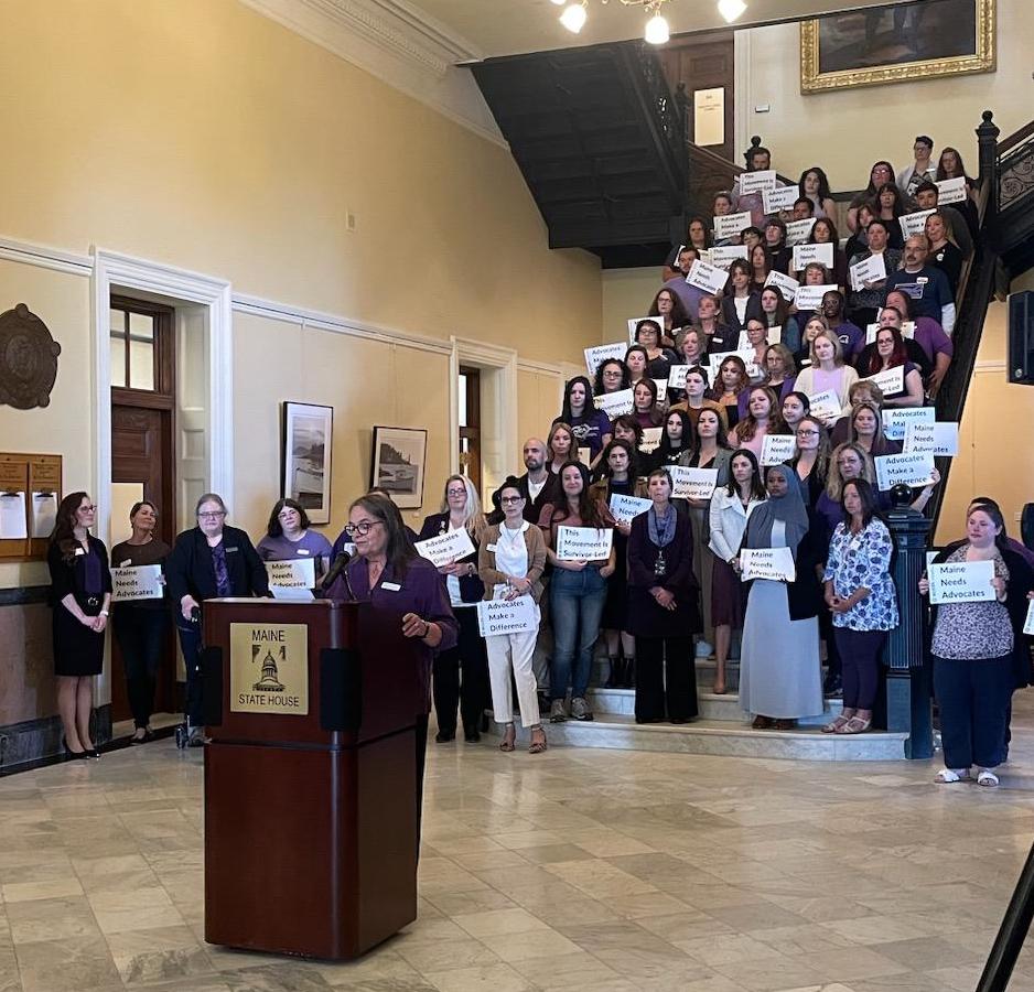 An image of people wearing purple clothing, standing on the stairs, behind a podium where a woman speaks. 